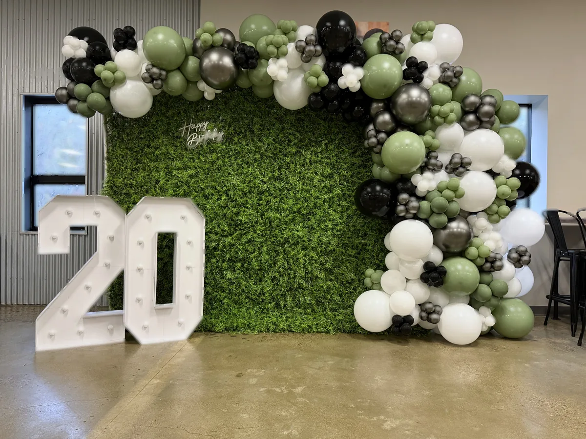 Sage green, black, and white organic balloon garland on a lush greenery wall with illuminated 20 marquee numbers and a Happy Birthday sign